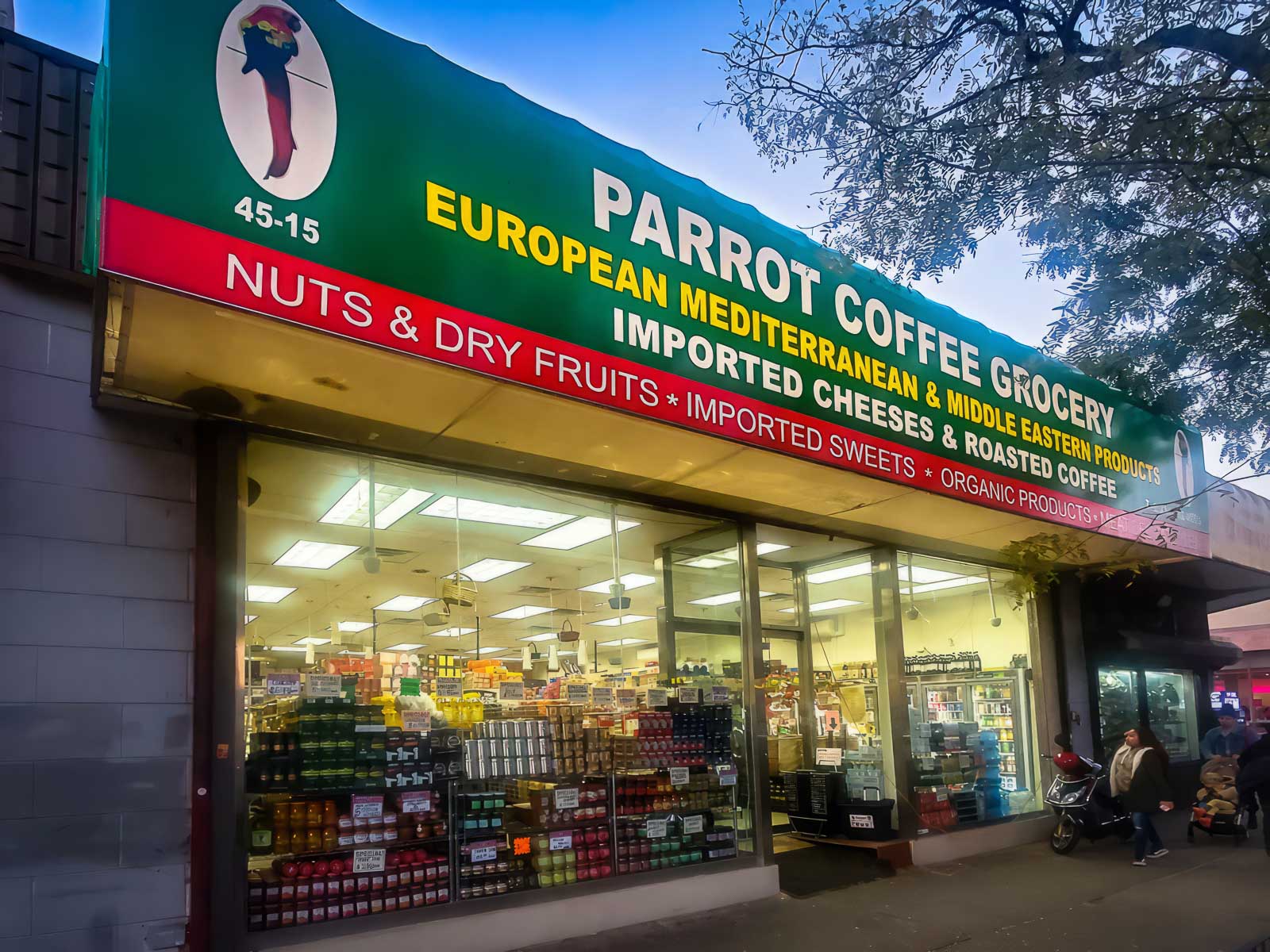 Illuminated storefront of Parrot Coffee Grocery at dusk, displaying a vibrant green awning with the logo of a parrot, advertising European Mediterranean & Middle Eastern products, nuts & dry fruits, imported cheeses, and roasted coffee.